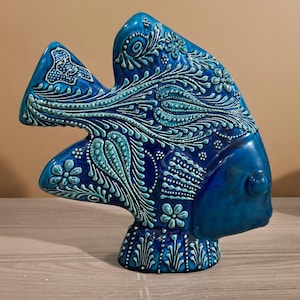 May include: A ceramic fish sculpture in shades of blue, with intricate floral and swirling patterns. The sculpture has a glossy finish and is approximately 12 inches tall. The design features a combination of light and dark blue hues, creating a visually appealing decorative piece.