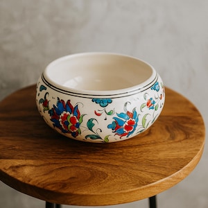 Könnte beinhalten: Eine weiße Keramikschale mit einem Blumenmuster in Rot, Blau und Grün. Die Schale hat einen schwarzen Rand und steht auf einem Holztisch. Die Schale hat einen Durchmesser von etwa 15 cm.