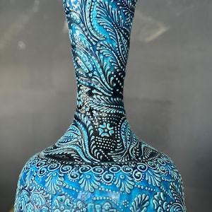 May include: A turquoise vase with detailed white floral patterns. The vase has a slender neck and a bulbous base. The intricate design covers the entire surface, creating a textured appearance. The vase's color is a vibrant turquoise, with white accents.