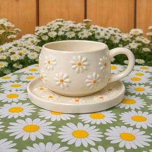 May include: Cream-colored ceramic teacup and saucer set with embossed daisy details. The teacup has a rounded shape with a handle. The saucer and teacup are decorated with white daisies with yellow centers. The set is on a green daisy-patterned tablecloth.