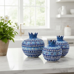 Puede incluir: Tres jarrones de cerámica azules y blancos con forma de granada y patrones intrincados. Los jarrones tienen una parte superior en forma de corona y están colocados sobre una encimera blanca. Una planta en maceta con flores blancas es visible en el fondo.