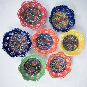 May include: A collection of seven ceramic bowls with scalloped edges and floral patterns. The bowls are in various colors, including red, blue, orange, yellow, and green. Each bowl features a unique, detailed design in the center.