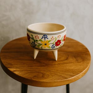 May include: A small, cream-colored ceramic bowl with a floral design in red, yellow, and blue. The bowl has three short, white legs and a black rim. The bowl is sitting on a round, wooden table.