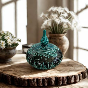 Może przedstawiać: Ozdobny, ręcznie malowany ceramiczny słoik z pokrywką. Słoik jest czarny z turkusowymi wzorami kwiatowymi i turkusowym zakończeniem. Słoik stoi na drewnianej powierzchni, z kwiatami w tle.