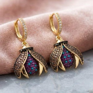 May include: A pair of gold hoop earrings featuring dangling insect-shaped charms. The charms have wings encrusted with brown crystals and a central body paved with vibrant magenta and turquoise stones, suspended from small gold hoops with clear crystals.