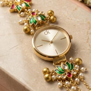 May include: A gold-tone analog watch with a round champagne dial featuring the ck logo. The watch is integrated into a decorative gold-tone chain bracelet adorned with green and red enamel lotus-shaped accents, small white beads, and dangling gold spheres.