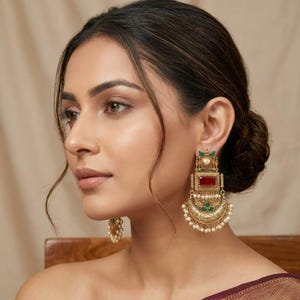 May include: Gold-toned earrings with a rectangular red stone and green accents. The earrings feature a half-moon design with small white pearls. The earrings are worn by a person with dark hair.