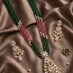 Pode incluir: Um conjunto elegante de colar e brincos com fios de contas verdes e rosa. O pingente e os brincos apresentam uma pedra rosa em forma de gota, rodeada por pequenas pedras brancas e pérolas. Também está presente um alfinete de flor dourado.