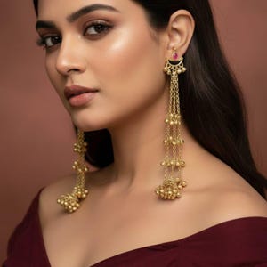 Puede incluir: Pendientes largos dorados con una parte superior en forma de media luna, una pequeña gema roja y múltiples cadenas con pequeñas cuentas redondas. Los pendientes tienen un diseño ornamentado y vintage. El fondo es marrón cálido.