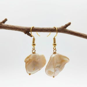 Peut inclure: Deux boucles d'oreilles pendantes dorées avec des pendentifs irréguliers en forme de coquillage, de couleur blanc cassé et nacré. Les boucles d'oreilles sont suspendues à une brindille brune sur fond blanc. Design simple et élégant.