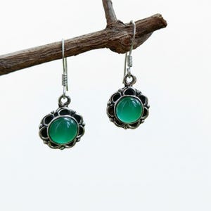 May include: A pair of silver-toned drop earrings with a round, bright green gemstone center. The earrings have a floral-shaped frame and are suspended from a simple hook. The earrings are displayed on a small, brown branch.