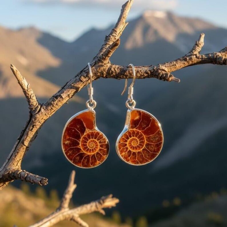 May include: A pair of silver-toned earrings featuring a fossilized ammonite design. The earrings have a spiral pattern in shades of brown and orange, set in a silver frame. They are suspended from a branch against a blurred mountain backdrop.