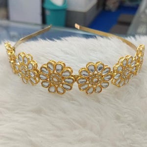 May include: A gold-toned headband featuring four flower-shaped embellishments. Each flower has a central cluster of small, clear stones surrounded by larger, clear stones, creating a sparkling effect. The headband is designed for a comfortable fit.