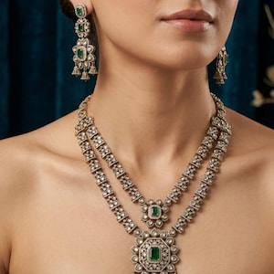 May include: A two-strand necklace and matching earrings set. The jewelry features a silver-toned metal with clear and emerald-green stones. The necklace has a floral pendant with a large emerald-green stone.