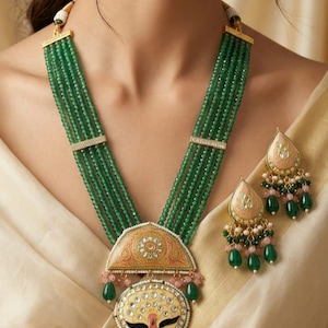 Pode incluir: Um colar de contas verdes e um conjunto de brincos a condizer. O colar apresenta vários fios de contas verdes, com detalhes dourados e um pendente com um design detalhado. Os brincos têm formato de gota com contas verdes.