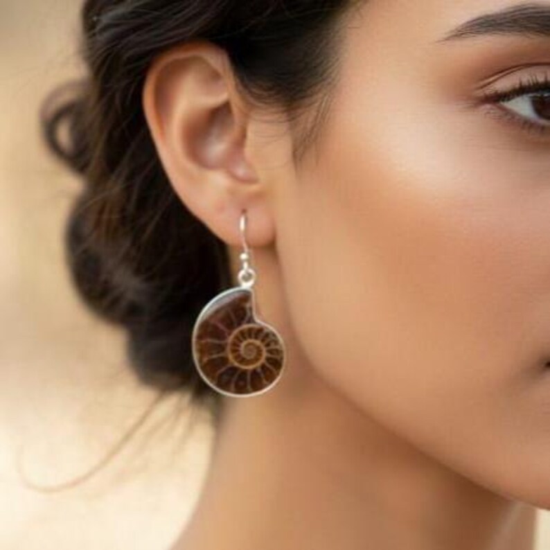 May include: A silver earring with a brown, spiral-shaped fossil pendant. The earring is a drop style, with a hook closure. The fossil has a detailed pattern and is set in a silver frame. The earring is worn on a person's ear.