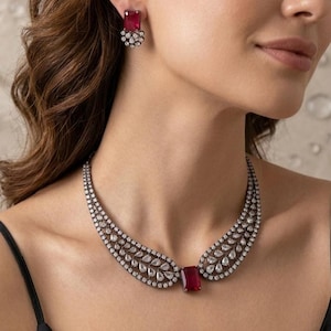 May include: A necklace and matching earrings set featuring a large, rectangular, red gemstone as a focal point. The necklace has a curved design with a series of smaller, clear gemstones. The earrings have a similar design. The jewelry is set against a neutral background.
