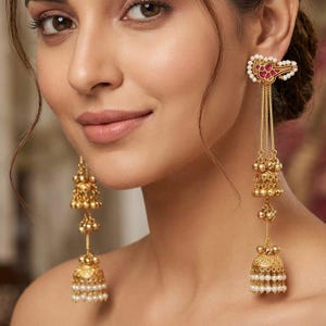 May include: Gold earrings with detailed patterns. One earring has a floral design with red accents and white pearls. The other has a long, dangling design with bell-shaped elements and small pearls.