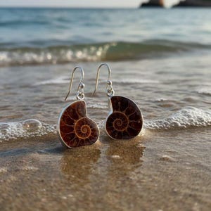 May include: A pair of silver earrings featuring a fossilized ammonite design. The earrings have a dark brown spiral pattern set in a silver frame. The earrings are displayed on a sandy beach with ocean waves in the background.