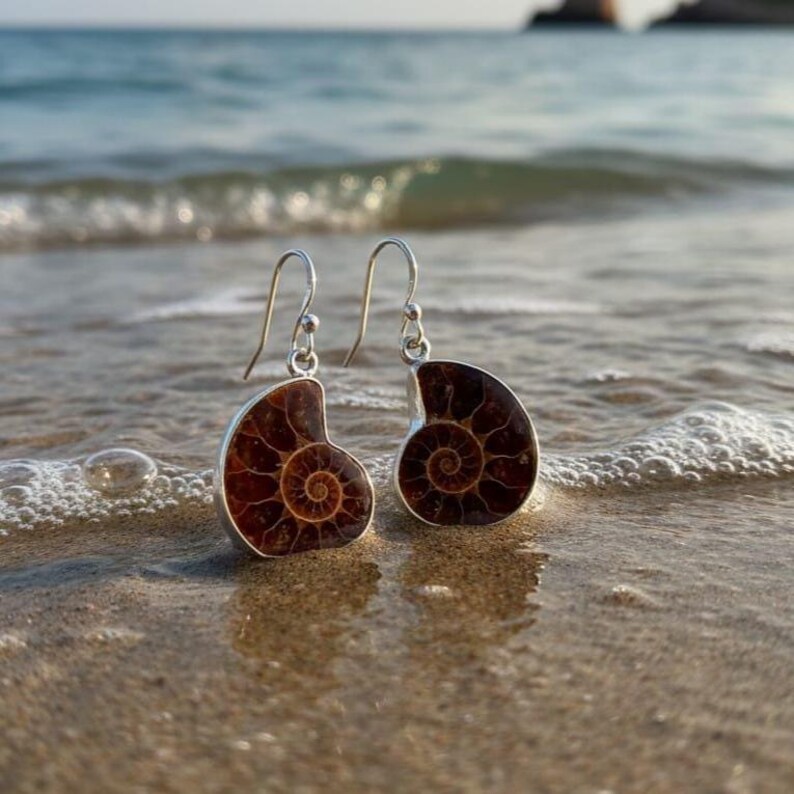 May include: A pair of silver earrings featuring a fossilized ammonite design. The earrings have a dark brown spiral pattern set in a silver frame. The earrings are displayed on a sandy beach with ocean waves in the background.