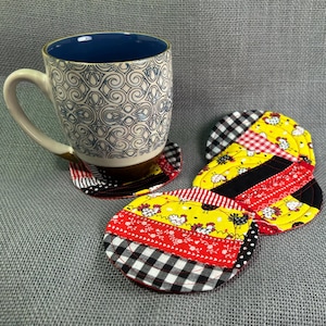 May include: A ceramic mug with a blue interior and a white and black patterned exterior sits on a matching coaster. Additional round coasters with patchwork designs in red, black, yellow, and white are also visible.