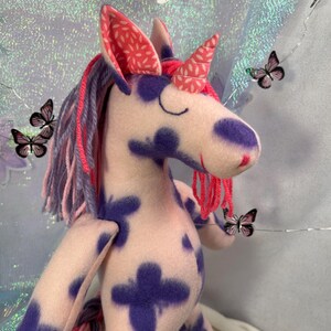 May include: A plush unicorn doll with a pink and purple color scheme. The unicorn has purple spots, a pink mane and tail, and a patterned horn and ears. Decorative butterflies and flowers are in the background.