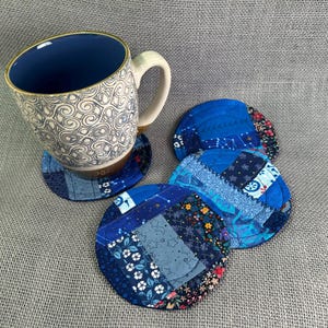 May include: A white mug with a blue interior and a decorative pattern sits on a blue patchwork coaster. Three additional blue patchwork coasters with floral and geometric patterns are also visible.