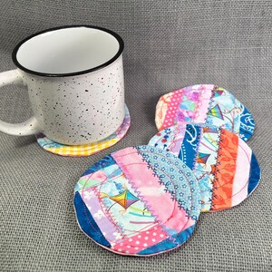 May include: A white speckled mug with a black rim sits on a colorful coaster. Several handmade fabric coasters with patchwork designs in pink, blue, and orange are also visible. The coasters feature kite patterns.