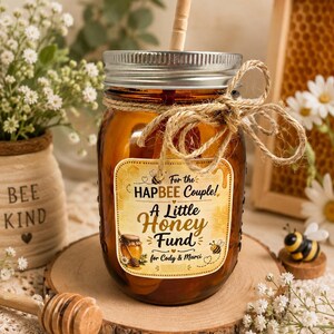 May include: An amber-colored glass mason jar with a silver metal lid and a wooden straw. A jute twine bow is tied around the neck. The label reads: For the HAPBEE Couple! A Little Honey Fund for Cody & Marci, featuring bee and honey pot graphics.