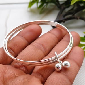 May include: A silver-colored bangle bracelet consisting of two thin, smooth, circular bands joined together. A small, dangling charm featuring two polished, spherical silver-colored beads hangs from the side of the double-banded bracelet structure.