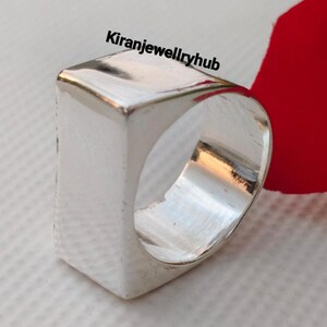 May include: A silver-colored, rectangular ring with a smooth, polished surface. The ring has a thick band and a flat, square top. The text "Kiranjewelleryhub" is visible on the top of the ring. The ring is set against a white background with a red accent.