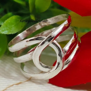 Peut inclure: Une bague en argent au design unique et entrelacé. La bague présente plusieurs bandes qui se bouclent et se croisent, créant une forme abstraite et accrocheuse. La bague est posée sur un fond rouge et vert.