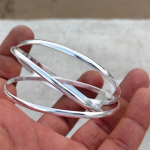 Könnte beinhalten: Ein Set aus drei ineinandergreifenden silberfarbenen Armreifen mit einer glatten, polierten und reflektierenden Oberfläche. Die dünnen, kreisförmigen Metallbänder sind in einem durchgehenden Schleifendesign verbunden und wirken geschichtet.