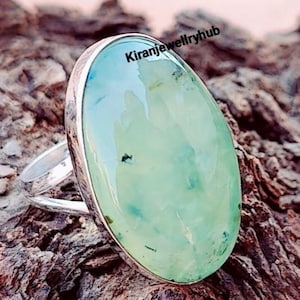 Könnte beinhalten: Ein silberner Ring mit einem großen, oval geschliffenen, hellgrünen Edelstein mit dunkleren grünen Einschlüssen. Der Ring liegt auf einer strukturierten, braunen Oberfläche. Der Text "Kiranjewelleryhub" ist sichtbar. Der Stein misst ca. 2,5 cm.
