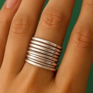 May include: A silver ring with a stacked band design. The ring features multiple thin, horizontal bands that create a layered effect. The ring is worn on a finger, showcasing its simple yet elegant style. The ring is made of a shiny, metallic material.