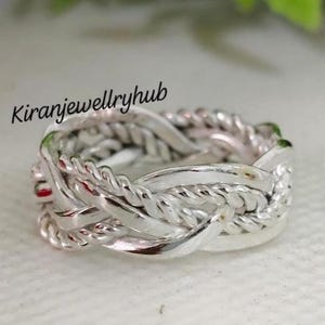 Puede incluir: Un anillo plateado con un diseño trenzado. El anillo presenta hebras entrelazadas, creando un patrón texturizado e intrincado. El texto "Kiranjewellryhub" es visible en la parte superior izquierda de la imagen.