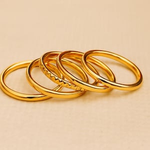 Pode incluir: Uma coleção de cinco anéis dourados. Um anel tem um design texturizado, enquanto os outros são lisos. Os anéis estão dispostos em uma linha ligeiramente curva, mostrando seu design simples, mas elegante. Os anéis são provavelmente de metal.