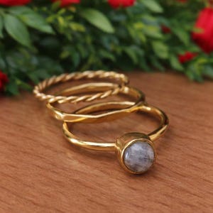 Op de afbeelding: Drie gouden ringen worden getoond op een houten oppervlak, met rode rozen op de achtergrond. De ringen omvatten een gedraaide band, een gladde band en een ring met een ronde, grijze edelsteen. De ringen hebben een warme goudkleur.