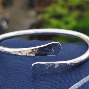 Puede incluir: Un brazalete plateado con textura martillada en los extremos abiertos. El brazalete se muestra sobre una superficie azul, con un fondo verde borroso.