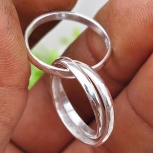 Peut inclure: Gros plan sur trois bagues argentées entrelacées. Les anneaux sont lisses et brillants, reflétant la lumière. Les bagues sont tenues dans une main, avec un arrière-plan vert flou.