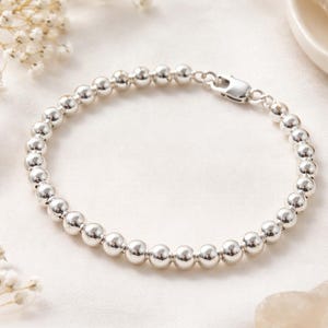 Pode incluir: Uma pulseira de contas prateadas com fecho seguro. A pulseira é composta por inúmeras contas pequenas, redondas e brilhantes. A joia é exibida sobre uma superfície branca texturizada, com iluminação suave e detalhes florais.