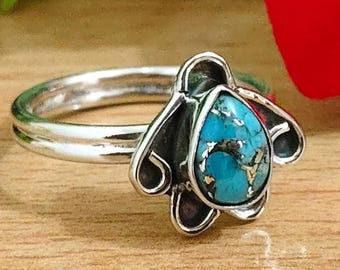 May include: A silver ring featuring a teardrop-shaped turquoise stone with gold flecks. The stone is set in a decorative silver bezel with a floral design. The ring is on a wooden surface.