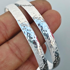 Op de afbeelding: Twee zilveren armbanden met een gehamerde textuur. De armbanden zijn rond en hebben een glanzend, reflecterend oppervlak. De afbeelding is een close-up, die de details van het metaalwerk laat zien. De armbanden worden op een hand getoond.