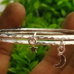 May include: Two silver bangles with a hammered texture. One bangle has a star charm, and the other has a crescent moon charm. The bangles are held against a background of green foliage.