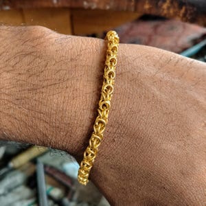 May include: A gold-coloured chain bracelet featuring a complex, interlocking Byzantine link pattern. The bracelet is constructed from numerous small, circular metal rings connected in a repeating, dense, and textured cylindrical design.
