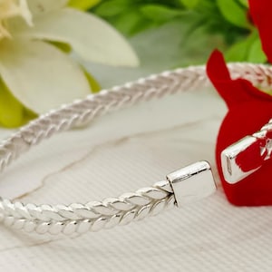 May include: A silver bracelet featuring a braided design. The bracelet has a clasp closure and is set against a floral backdrop. The braided pattern gives the bracelet a textured appearance.