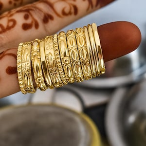 May include: A collection of ten gold-toned metal rings stacked together. Each ring features a distinct surface texture, including geometric patterns, floral-inspired motifs, smooth polished bands, and repeating oval shapes in a bright finish.