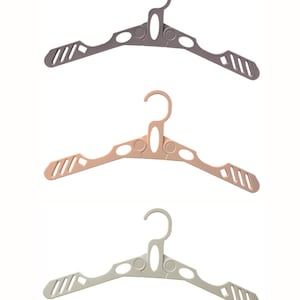 Puede incluir: Tres perchas de plástico en tonos gris, melocotón y verde claro. Cada percha tiene un gancho curvo, un soporte central y ranuras anguladas en los brazos. Las perchas están diseñadas para guardar ropa.