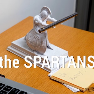Puede incluir: Un portalápices de estatua espartana blanca con un bolígrafo negro. La estatua está arrodillada con un escudo y un casco. El texto "the SPARTANS" está en la parte inferior. Una pila de notas adhesivas amarillas con "Be Happy!" está a la derecha.
