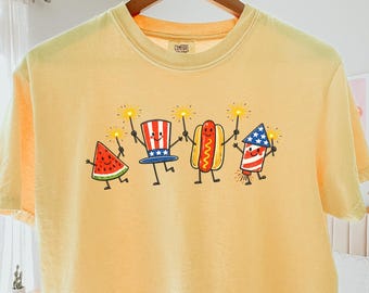 May include: A pale yellow short-sleeve t-shirt featuring a graphic print of a smiling watermelon slice, a top hat, a hot dog, and a firework, all holding sparklers. The shirt has a crew neckline and a boxy fit with a Comfort Colors brand label.